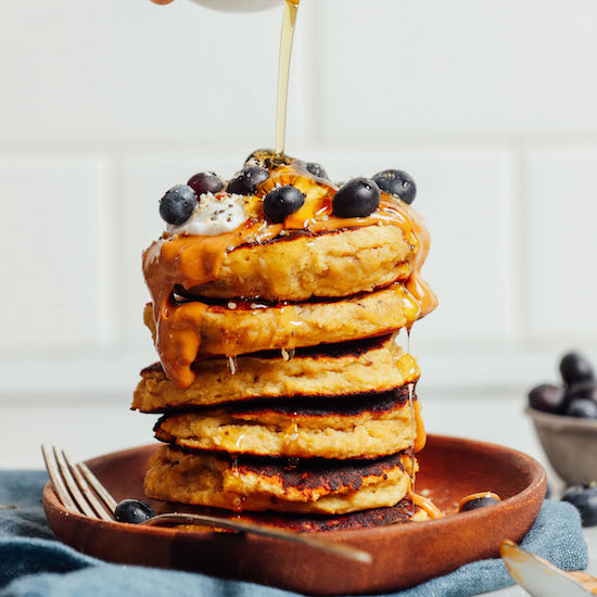 Drizzling syrup onto a stack of Banana Egg Pancakes topped with fresh blueberries and peanut butter