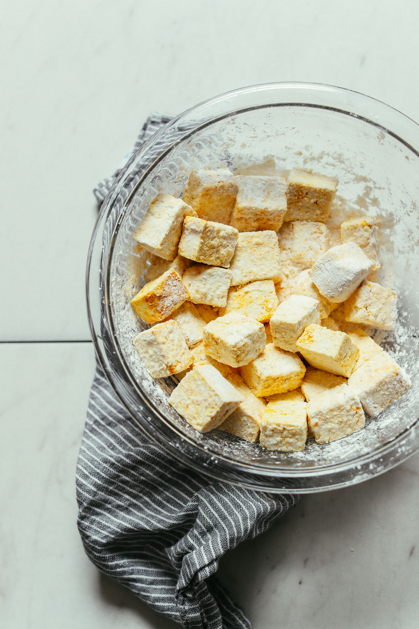 Overhead image of tofu tossed in spices and cornstarch in a glass mixing bowl with striped blue towel underneath