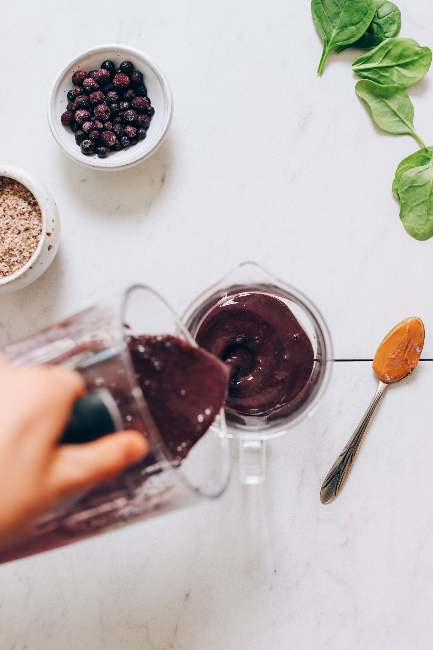 Overhead image of smoothie being poured into a glass measuring cup with spinach, wild blueberries, and a spoonful of peanut butter on the side.