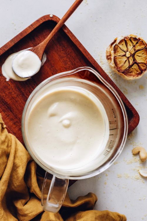 Spoon and glass of vegan bechamel sauce next to a head of roasted garlic