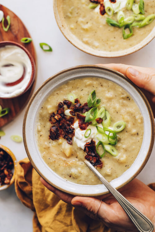 Holding a bowl of potato leek soup topped with green onion, coconut bacon, and sour cream