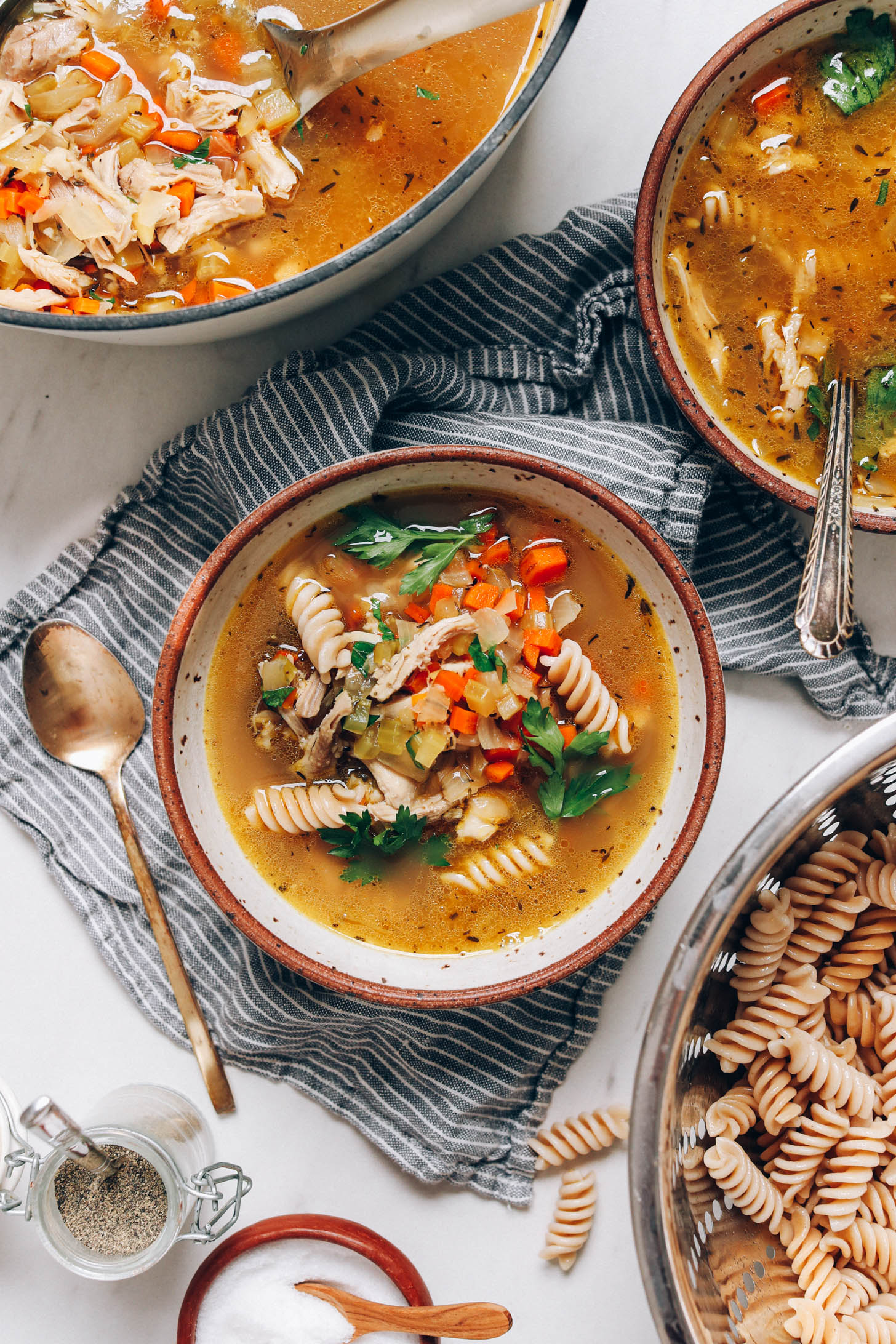 Colander of gluten-free noodles next to bowls and a Dutch oven of chicken noodle soup