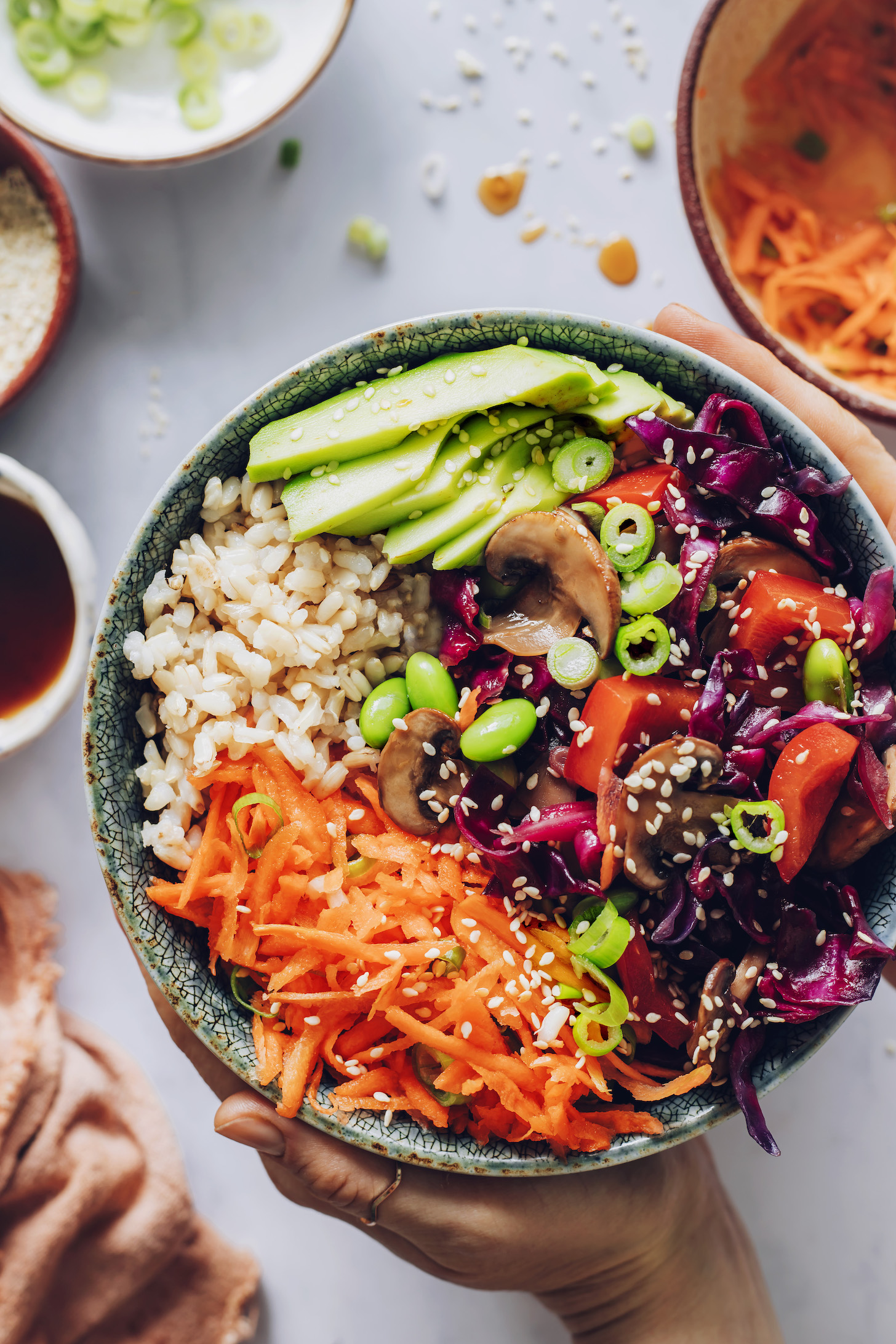Holding a vegan bowl of brown rice topped with avocado, carrot slaw, and sautéed veggies and edamame cooked in teriyaki sauce