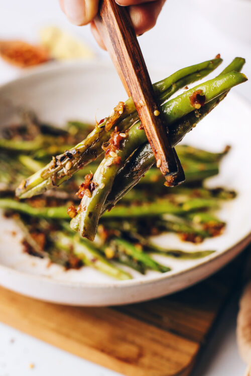 Chopsticks holding spicy stir-fried green beans in front of a bowl