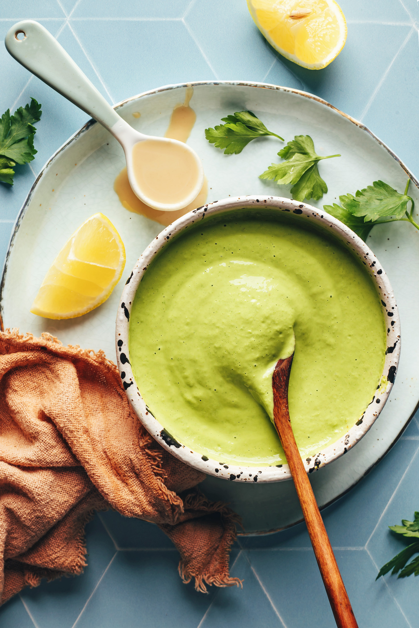 Bowl of herby green tahini sauce next to lemon, parsley, and a spoonful of tahini